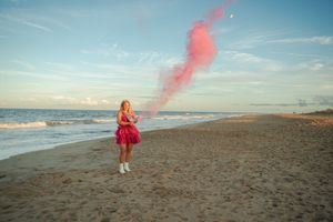 A person in a red dress stands on a beach holding a pink smoke bomb against a blue sky and ocean backdrop.