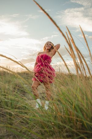 A figure in a flowing pink dress stands among tall beach grass at sunset with a dreamy summer atmosphere.
