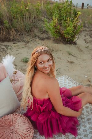 A person in a flowing magenta dress poses gracefully on a sandy beach with coastal vegetation in the background.