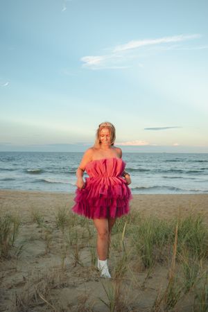 A person in a bright pink tulle dress poses on a beach at sunset wearing white boots.