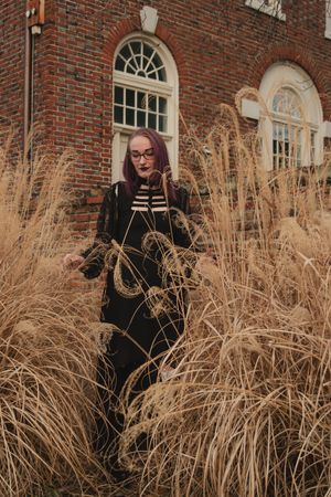 A person in black attire poses against a brick building with ornate windows and dried ornamental grasses in the foreground.
