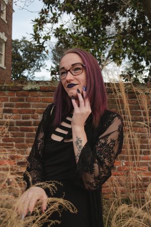 Person with burgundy hair and glasses laughs while standing against a rustic brick wall with dried grasses in background.