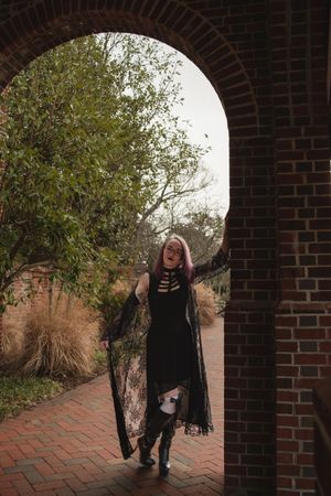 A figure in black gothic attire poses dramatically in a brick archway on an autumn day.