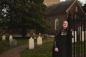 Gothic figure stands near historic church buildings and gravestones at dusk.