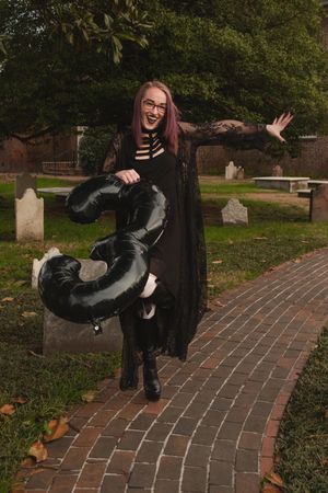 Someone in a black dress and boots dances playfully on a brick pathway in a cemetery during autumn.