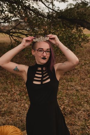 A person in a black sleeveless top adjusts a tiara while posing outdoors in an autumn setting.