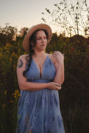A person in a flowing blue dress and wide-brimmed hat poses with eyes closed at sunset among wildflowers.