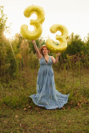 A person in a flowing blue dress holds large gold number 33 balloons in a sunlit outdoor field at sunset.
