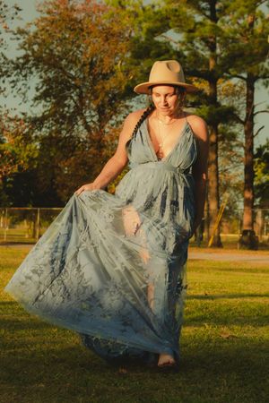 A person in a flowing blue dress and sun hat twirls gracefully in a sunlit park during golden hour.