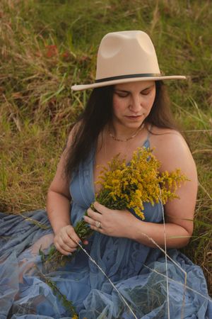Person in a flowing blue dress and sun hat sitting in tall grass with yellow wildflowers.