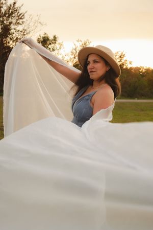 A flowing white veil billows in the golden sunset light as someone in a denim top and hat poses outdoors.
