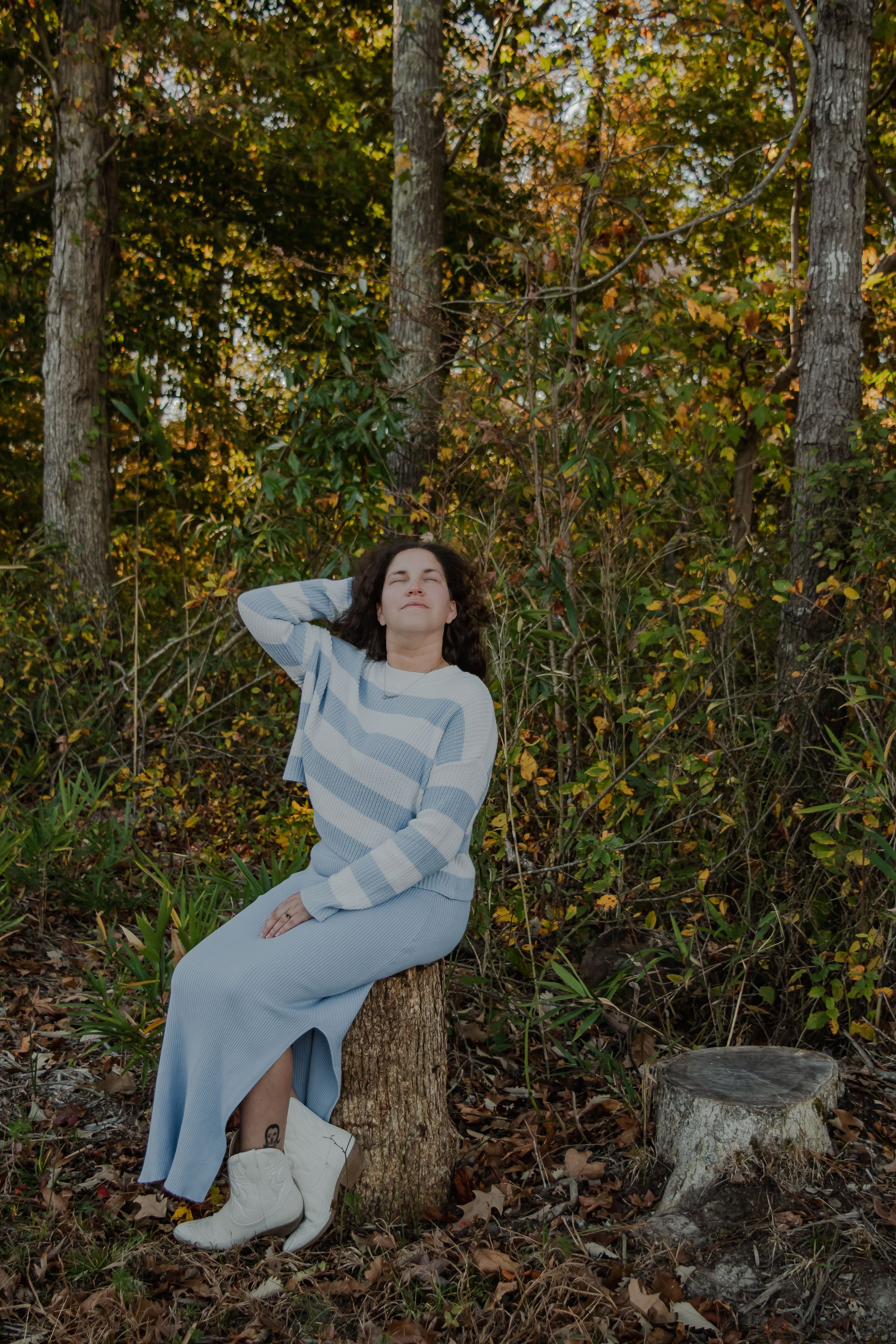 A person in a light-colored outfit reclines against a tree trunk in an autumn forest setting with golden foliage.
