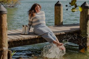 Someone in a cozy gray sweater sits on a wooden dock splashing their feet in the water on a sunny autumn day.