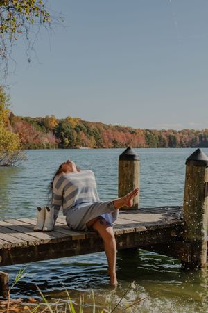 Person lying on wooden dock splashing water with their feet at a scenic lake surrounded by autumn foliage.