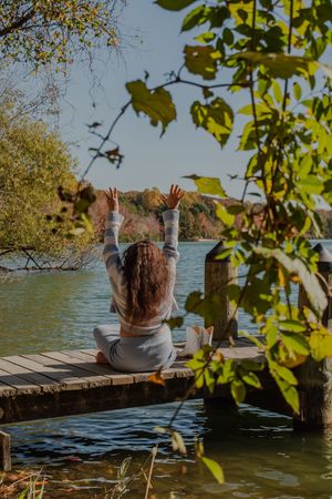 A person sits peacefully on a wooden dock by a tranquil lake surrounded by autumn foliage on a sunny day.