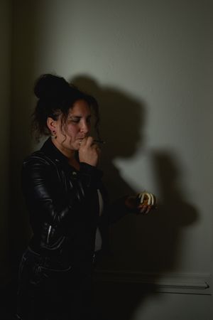 A person in a black leather jacket stands thoughtfully against a dark wall casting dramatic shadows.