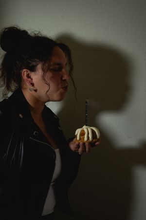 A person in a black jacket leans towards a lit birthday candle casting dramatic shadows against a dark background.