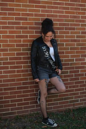 Person in black jacket and fishnet stockings sits against red brick wall wearing casual sneakers.