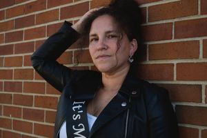 A person in a black jacket leans against a brick wall while looking off to the side.