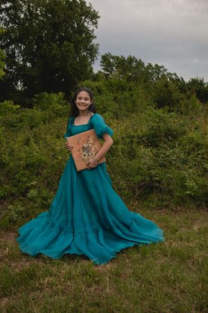 Someone poses outdoors in a flowing teal dress with short sleeves against a backdrop of green foliage.