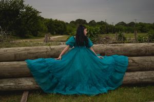 A flowing teal dress spreads dramatically across wooden logs in a rustic outdoor setting.