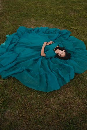 A person in a flowing teal dress lies gracefully on green grass in an artistic outdoor photo series.