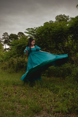 A figure in a flowing teal dress spins gracefully in a grassy field with trees in the background.