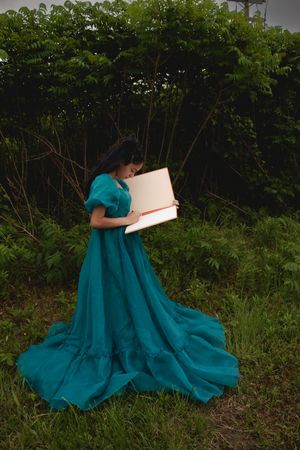 A person in a flowing teal dress sits reading a book outdoors against a backdrop of lush green foliage.