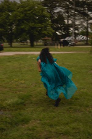 A person in a flowing teal dress spins and twirls gracefully across a grassy field on a cloudy day.