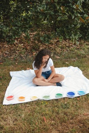 A white blanket laid on grass with rainbow colored paint dots arranged in a spectrum while someone sits in contemplation.