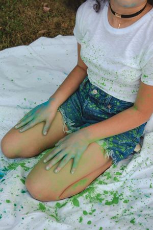 Person sitting on white sheet wearing denim shorts with green paint splattered on hands and clothing during messy art session.