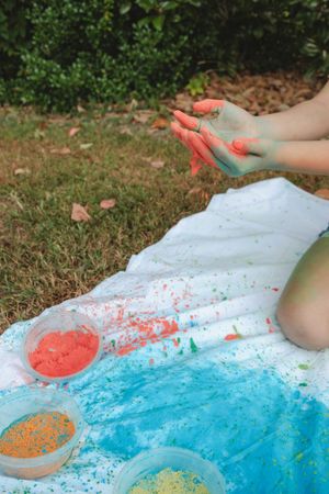 Hands play with colorful powder paint on a white sheet outdoors with blue, orange and green pigments scattered about.