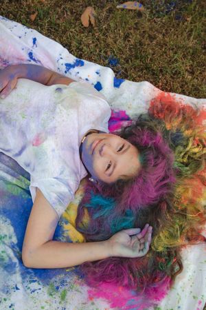 Someone lies on grass with vibrant powder paint colors covering their white clothes and hair in a rainbow pattern.