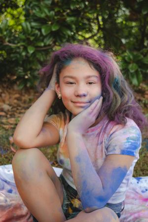 A person with colorful purple and pink hair poses playfully outdoors against a green leafy background.