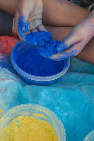 Hands scooping bright blue powder paint from a container with yellow paint visible in the background.