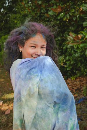 A sequence of photos showing someone playfully hiding behind a blue and white tie-dye fabric outdoors among green foliage.