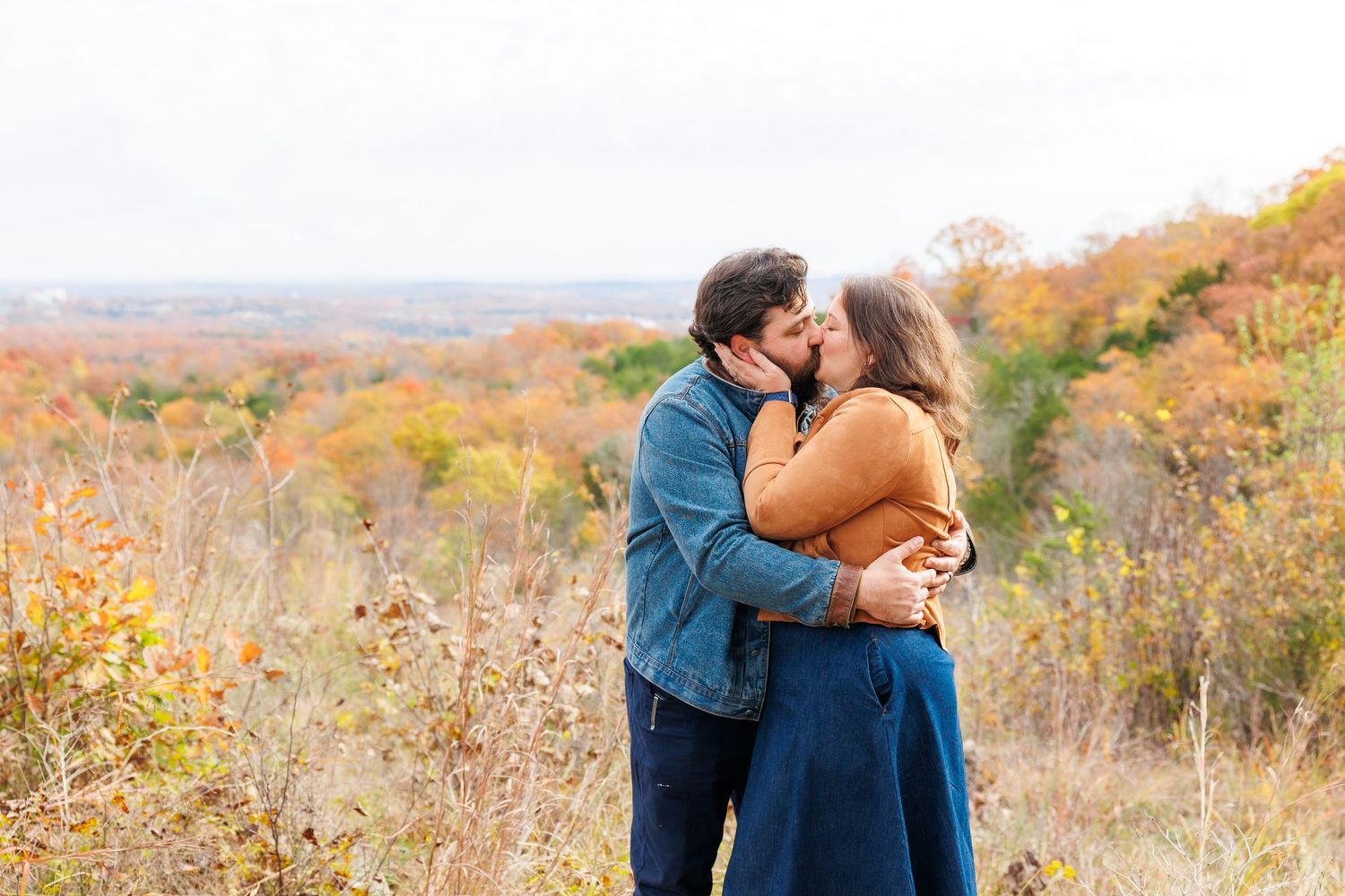 A couple stands together by a serene lake surrounded by autumn trees and mountains in the background.