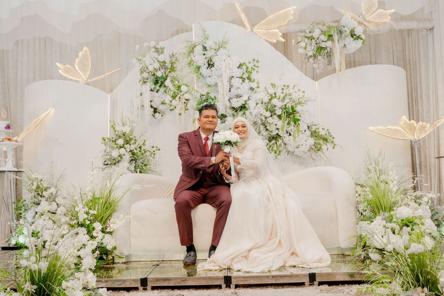 Newlyweds in burgundy suit and white dress sit on decorated platform with butterfly accents.