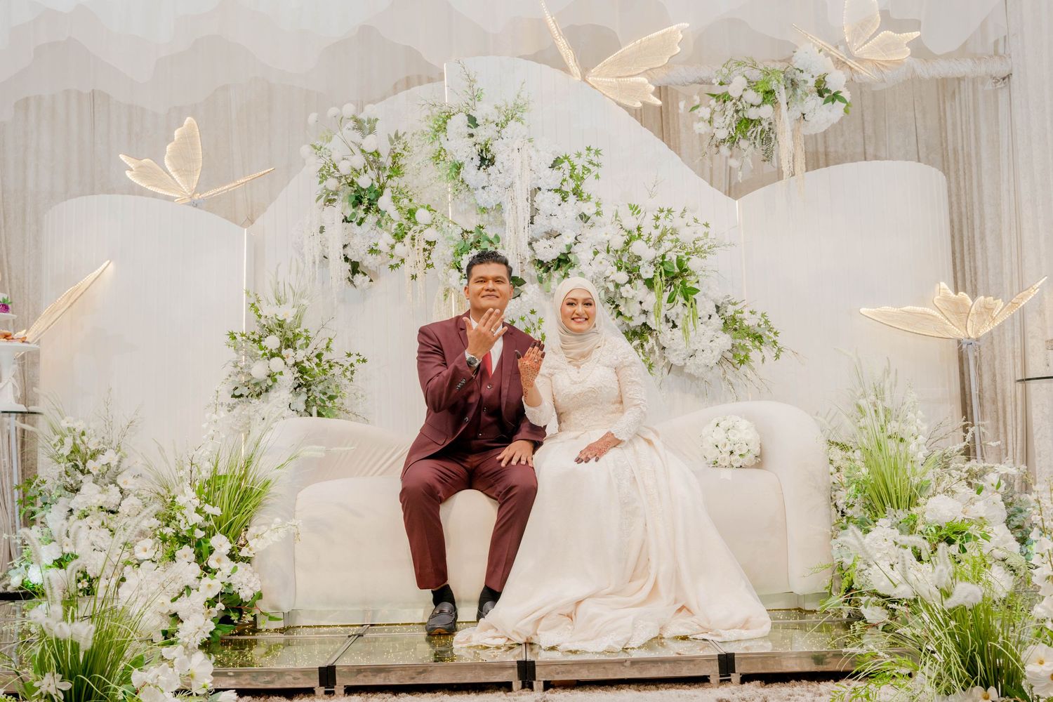 Bride in white dress and groom in burgundy suit pose on elegant white stage with greenery.