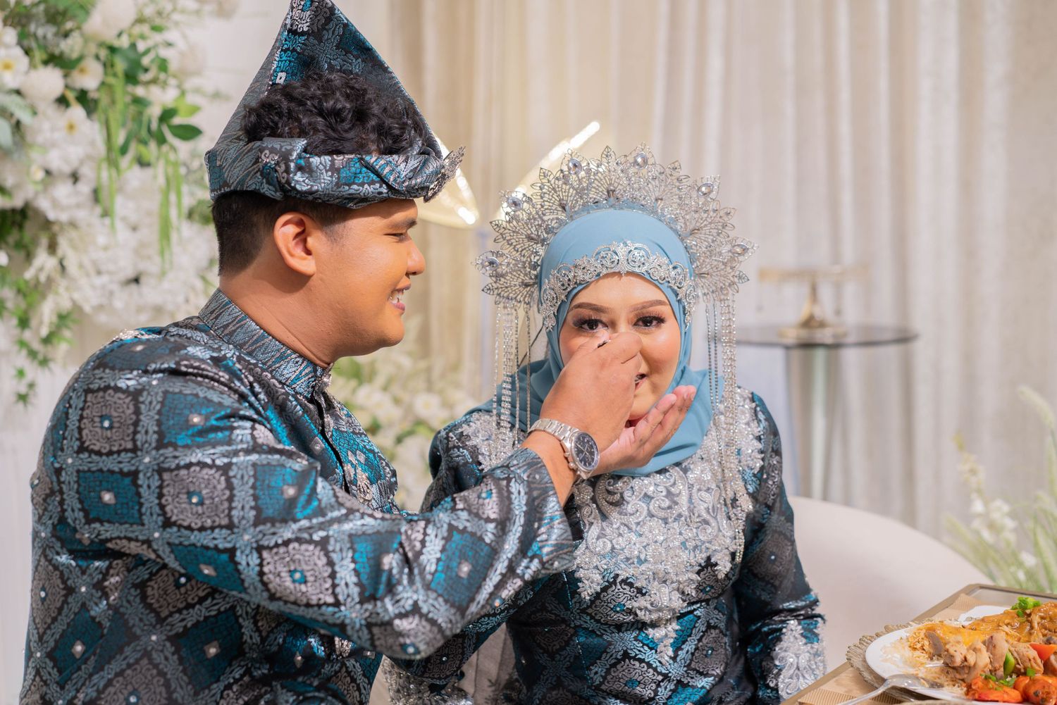 A couple in matching blue traditional attire feeding each other during a celebration.