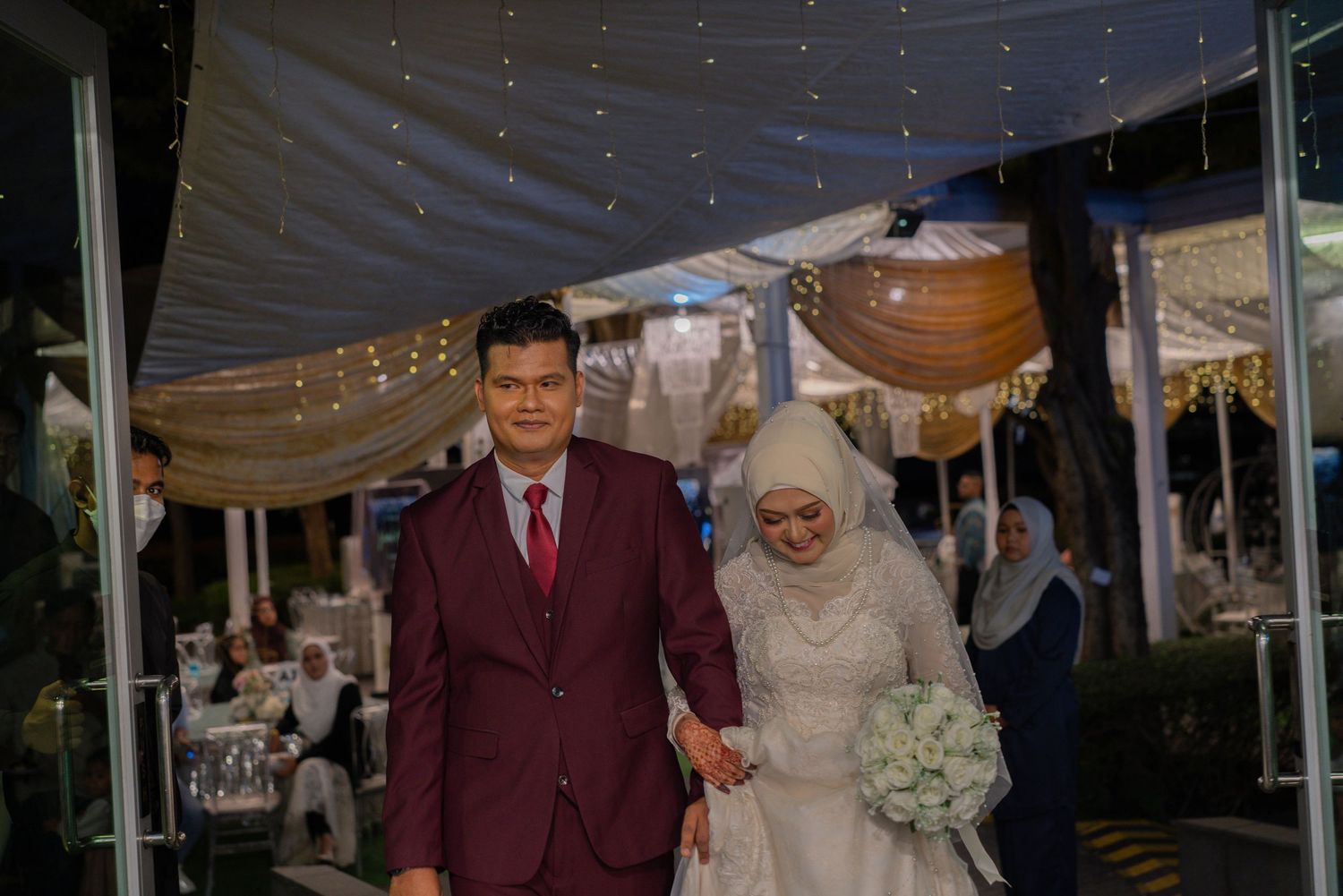 A couple in burgundy suit and white hijab dress walking during their wedding celebration.