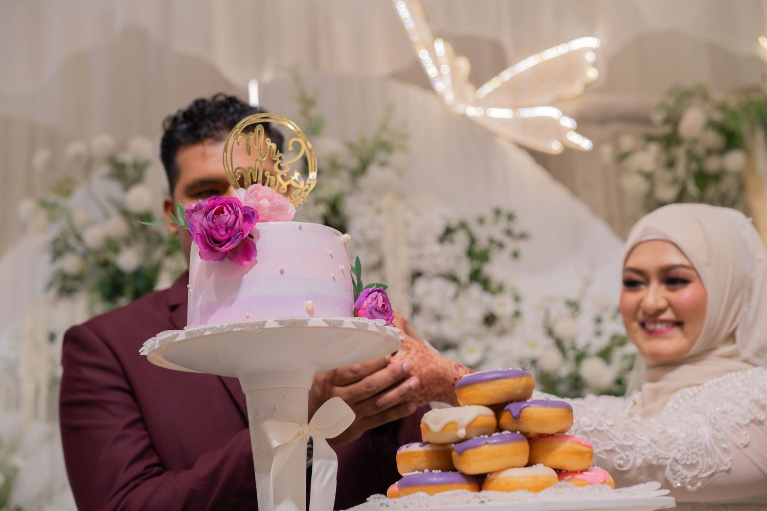 A wedding cake and donuts display with decorative butterfly lights in the background.
