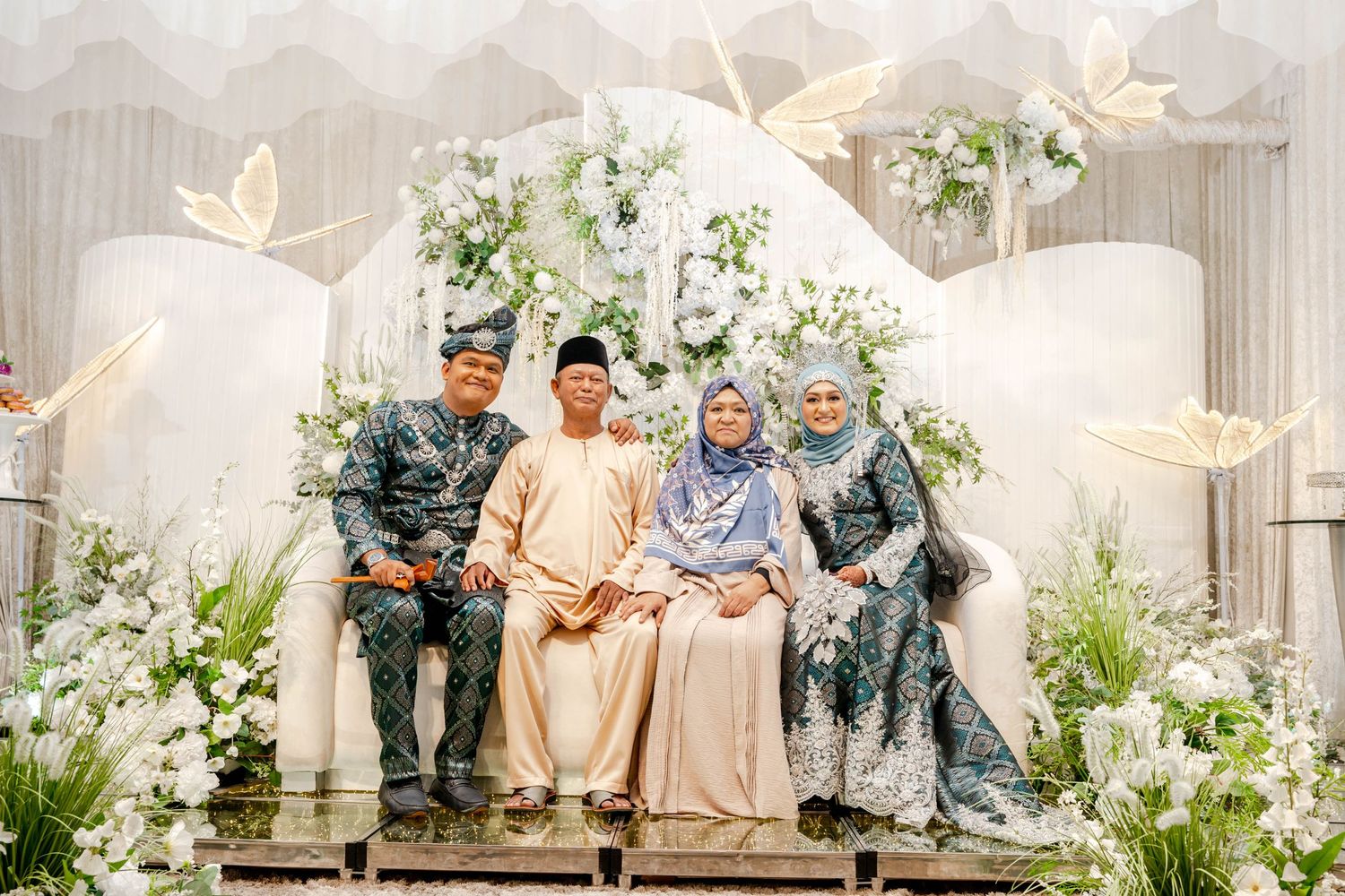 A group in traditional Malay attire seated on a decorated stage with butterfly motifs.