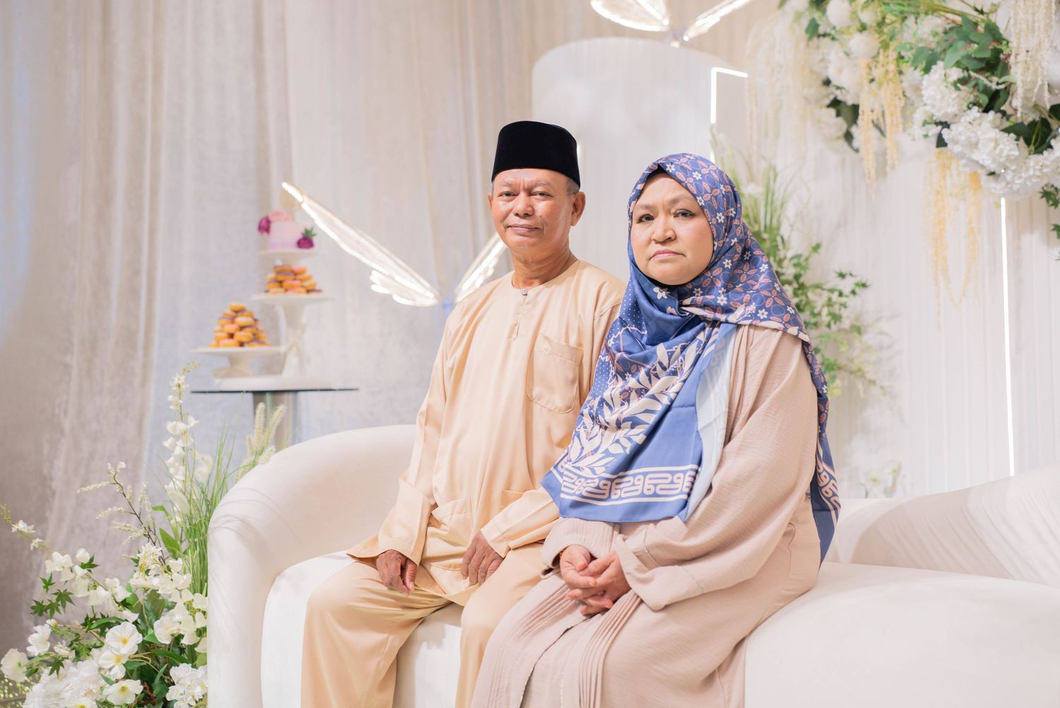 A couple in traditional Muslim attire seated on an elegant white couch with floral decorations.