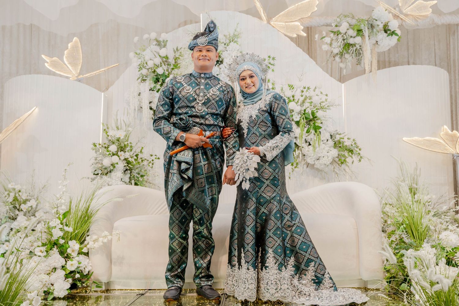 Couple in traditional Indonesian wedding attire pose against elegant white floral backdrop.