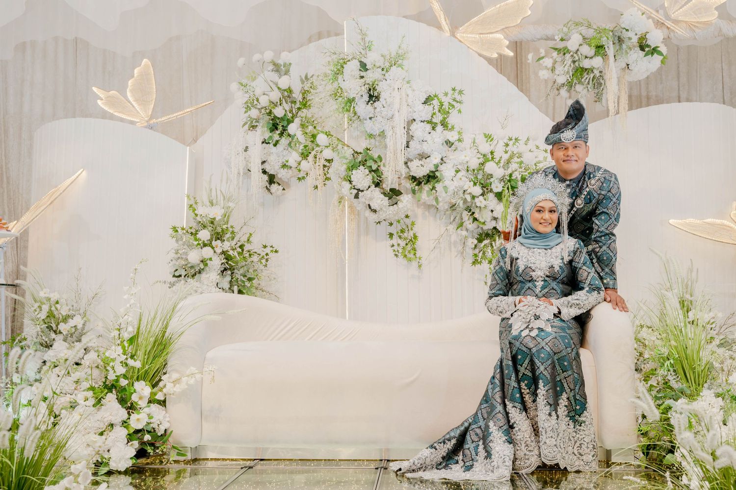 Wedding couple in matching blue traditional outfits sit together on white stage with floral decor.