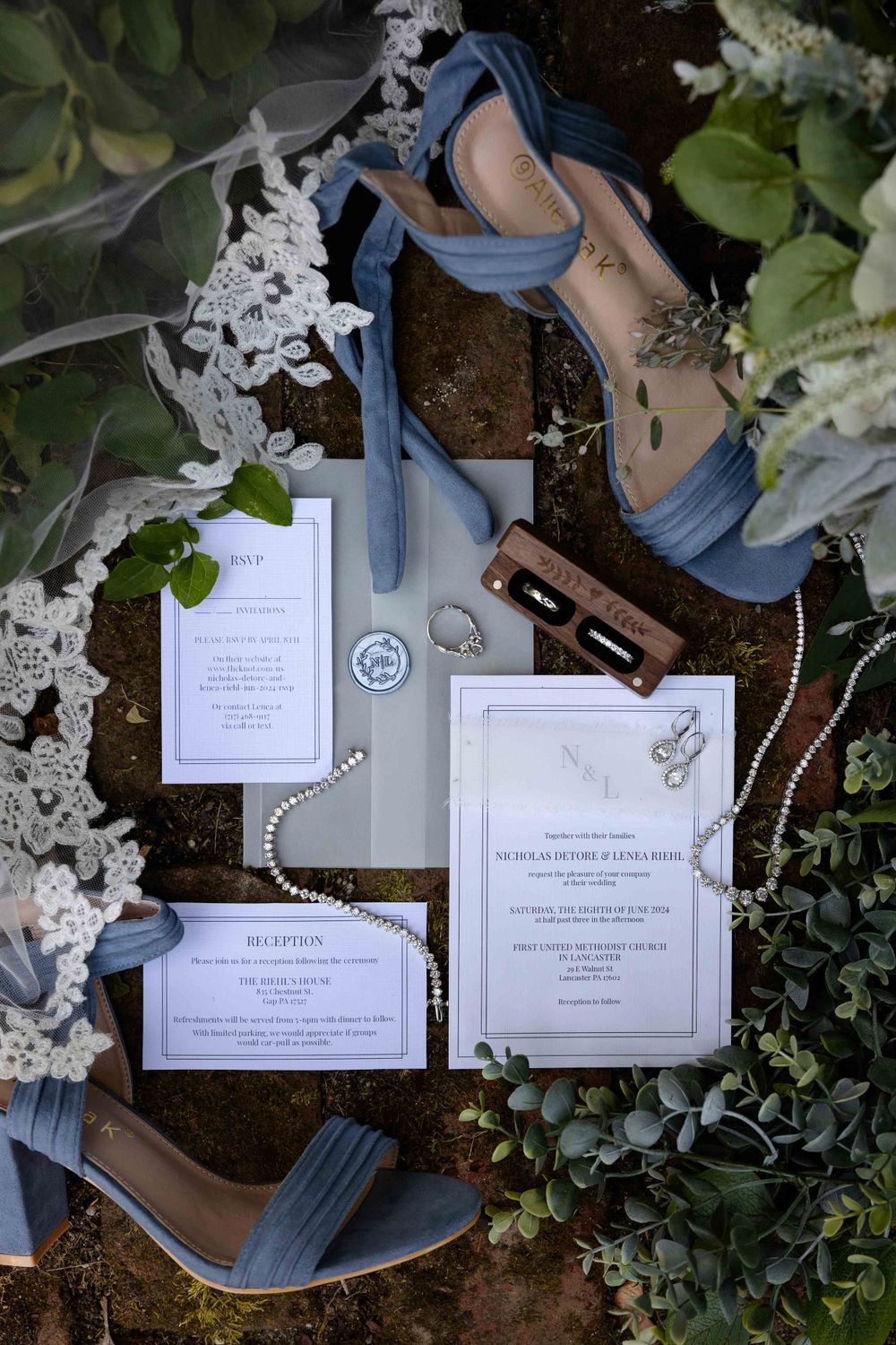 Wedding invitation suite with blue heels and white floral decor arranged on dark background.