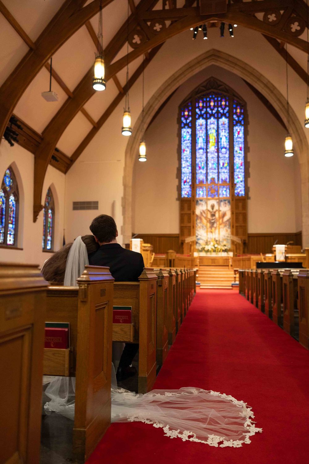 White wedding veil drapes across red carpet aisle in a church with stained glass windows and wooden pews.