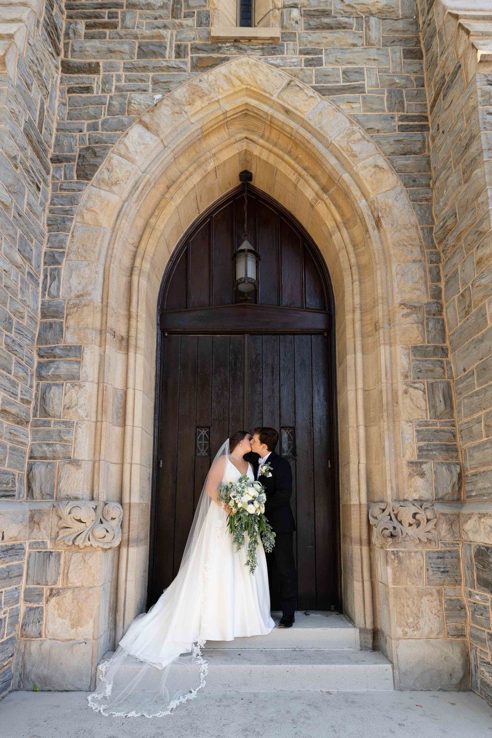 A bride and groom share a romantic moment under a grand Gothic stone archway entrance of a church.
