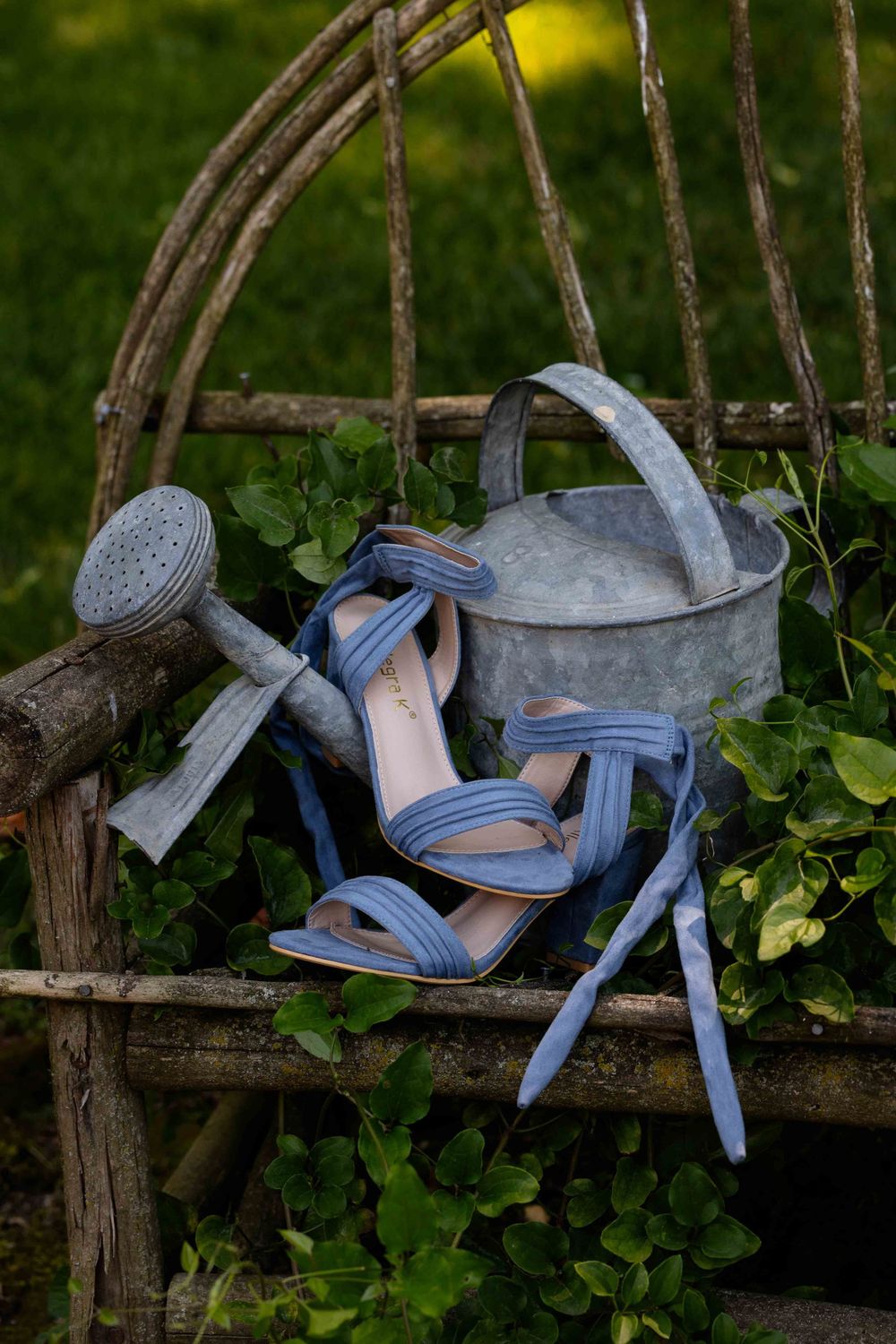 Blue sandals and a metal watering can sit on rustic wooden garden furniture surrounded by green foliage.